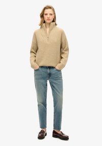 Suéter de punto texturizado beige con cremallera de un cuarto y un cuello ancho, combinado con jeans azules deslavados y mocasines burdeos. Fondo liso.