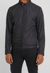 Svart jacka med rutmönster, hög krage, framdragkedja och två sidofickor med dragkedja. Tillverkad av ett mjukt och lätt material.