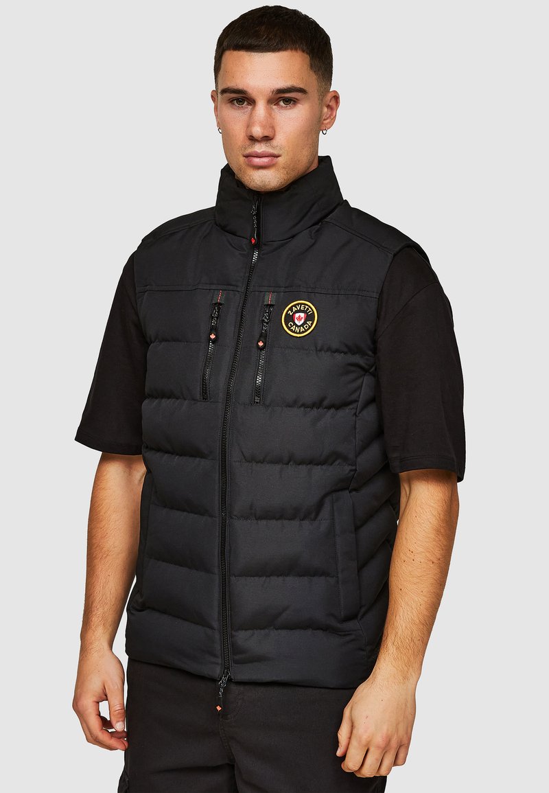Zwarte gewatteerde vest met een hoge kraag, ritssluiting aan de voorkant, twee borstzakken en een rond logo-embleem. Mouwloos ontwerp, matte textuur.