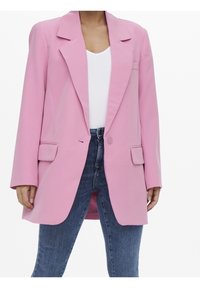 Donna che indossa un blazer oversized rosa pastello con tasche a patta, una maglietta bianca a V e jeans blu a vita alta, su uno sfondo bianco.