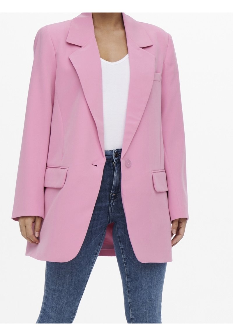 Donna che indossa un blazer oversized rosa pastello con tasche a patta, una maglietta bianca a V e jeans blu a vita alta, su uno sfondo bianco.