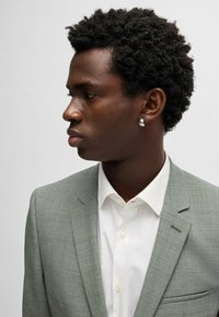 Veste de costume tailleur gris clair avec une finition texturée, associée à une chemise habillée blanche. Présente une boucle d'oreille et des cheveux courts et bouclés.