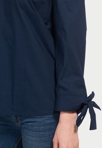 Marineblaue Bluse mit langen Ärmeln und gebundenen Manschetten. Hergestellt aus glattem Stoff, hat sie eine lockere Passform und ein einfaches, minimalistisches Design.