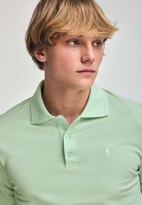 Ljusgrön polo skjorta med krage, texturerad tyg, tre knappar och en liten broderad logotyp på vänster bröstområde.