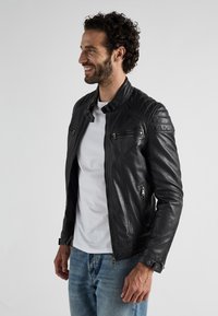 Zwarte leren motorjack met gestikte accenten op de schouders, voorzakken met rits en een kraag met drukknoop. Gestyled met een wit T-shirt.
