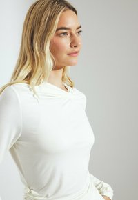 Blonde woman with light blue eyes wearing a long-sleeve white top with gathered details at shoulder and waist, facing right.
