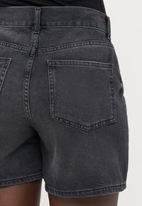 Shorts de mezclilla en negro desgastado, con un diseño de cintura alta, dos bolsillos traseros y costuras en contraste. La textura es suave con un ligero estiramiento.