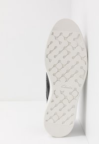 Chaussure en cuir noir avec une tige texturée, dotée d'une semelle en caoutchouc blanche à motif ondulé et de la marque "Clarks" sur la semelle.