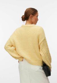 Femme aux cheveux bruns attachés en chignon, portant un pull jaune épais en tricot et une jupe blanche, tenant une pochette noire, de profil sur un fond uni.