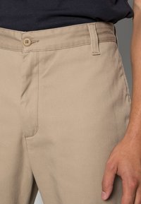 Calças de algodão bege com fecho de botão, bolsos laterais e uma textura suave. Apresentam um corte reto e costuras robustas.