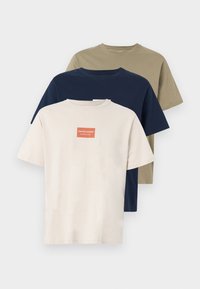 Τρία μονόχρωμα T-shirts με στρογγυλή λαιμόκοψη σε μπεζ, ναυτικό μπλε και ελιά, με το μπεζ μπλουζάκι να φέρει ένα μικρό κόκκινο λογότυπο στο στήθος.