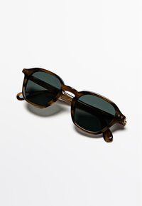Hnědé sluneční brýle v želvovinovém vzoru s tmavě zelenými čočkami. Čtvercové obroučky s jemnými detaily, tenké ramínka a dvě malé nýty na každé straně.