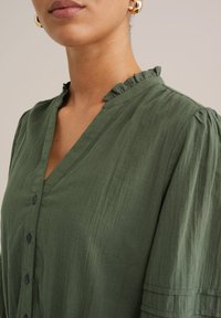 Blusa verde oliva con scollatura a V, colletto arricciato e dettagli con bottoni. Realizzata in tessuto texturizzato con pieghe sottili sulle spalle.