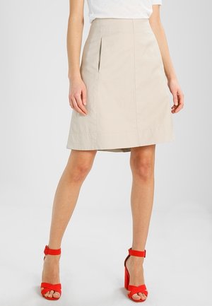Kvinde iført beige knælængde nederdel og røde høje hælede sandaler, stående mod en ensfarvet lys baggrund.