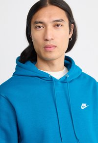Jaunas žmogus su tiesiais ilgais tamsiais plaukais, dėvintis sidabrinius auskarus ir mėlyną Nike džemperį prieš balto fono.