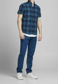 Man som bär en kortärmad blå och svart rutig skjorta, vit t-shirt under, blå jeans och vita sneakers, stående mot en enfärgad bakgrund.