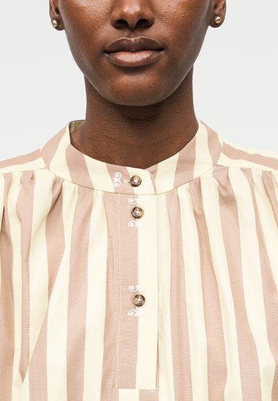 Άτομο με καφέ επιδερμίδα που φοράει μπεζ και κρεμ πουκάμισο με κάθετες ρίγες, μικρά στρογγυλά κουμπιά και χρυσά σκουλαρίκια με καρφάκια, πάνω σε λευκό φόντο.