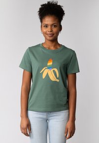 T-shirt en coton vert avec un grand motif de banane jaune surmonté de rayures arc-en-ciel. Manches courtes, col rond, coupe décontractée.