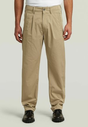 Pantalon beige ample taille mi-haute avec plis devant, porté avec des chaussures noires à lacets et un haut blanc, visible de la taille aux pieds.