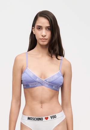 Un mannequin aux longs cheveux foncés porte un bralette lilas Moschino et une culotte blanche avec une ceinture ornée d’un cœur Moschino et du mot « You », sur un fond uni.