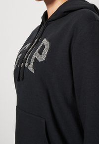 Felpa nera con cappuccio e tasca frontale, caratterizzata da un logo "GAP" in argento glitterato. Ha un cappuccio con cordini e un tessuto morbido e strutturato.