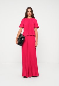 Ein heißpinkfarbener, gefalteter Oberteil und eine Kombination aus weit geschnittenen Hosen, ausgestattet mit kurzen, ausladenden Ärmeln. Begleitet von einer schwarzen, glänzenden Clutch.