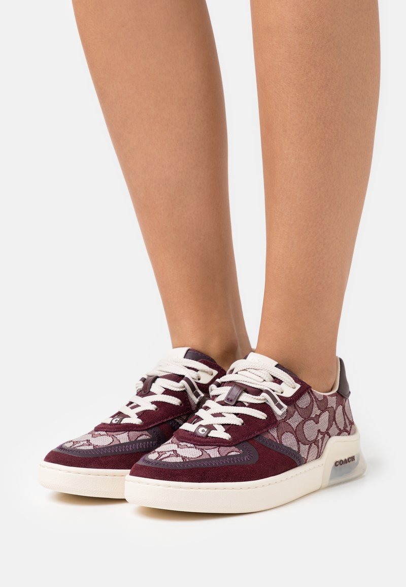 Jambes portant des baskets Coach bordeaux et rose clair avec des lacets blancs et des semelles blanches, présentées sur un fond blanc uni.