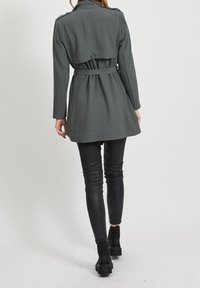 Donna che indossa un trench grigio scuro con cintura, pantaloni in pelle nera e stivali neri alla caviglia, in piedi su uno sfondo chiaro e semplice.