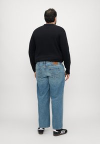 Maglietta nera a maniche lunghe, jeans azzurri larghi con una toppa in pelle marrone e sneakers nere con strisce bianche, viste da dietro.