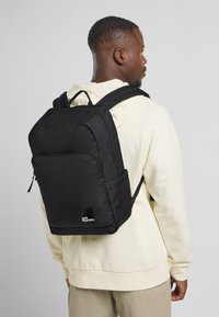 Mochila negra con una estructura acolchada, bolsillo frontal con cremallera, bolsillos laterales de malla y una etiqueta de marca. Lleva puesta una sudadera ligera.
