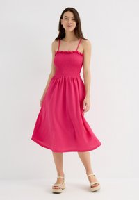Vestido midi rosa con el cuerpo fruncido, tirantes finos y escote con volantes. Tela suave con un acabado texturizado, silueta fluida.