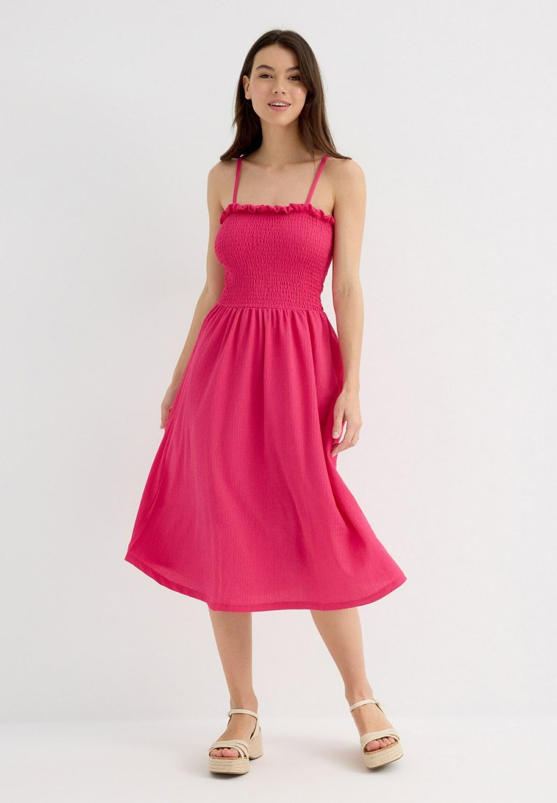 Vestido midi rosa con el cuerpo fruncido, tirantes finos y escote con volantes. Tela suave con un acabado texturizado, silueta fluida.