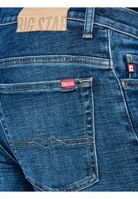 Aizmugurējā kabata zilajām džinsa biksēm ar dzeltenām šuvēm un divām sarkanām Big Star zīmola birkām uz kabatas un jostas vietā.