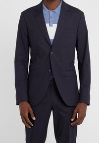 Marinblå kavaj med två knappar, slagkrage och sidofickor. Parat med en ljusblårandig polo skjorta. Slät tygtextur.