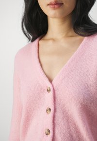 Ljusrosa kofta i mjukt, strukturerat tyg med V-ringning och fem träknappar fram. Korta ärmar.