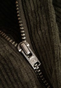 Tessuto di corduroy verde con una cerniera metallica visibile. La cerniera presenta un tirante argentato e una superficie testurizzata e rigata.