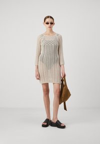 Knit ruha nyitott szövési mintával krémszínben. Háromnegyedes ujjú, testhez simuló sziluett. A modellen barna táska és fekete szandál található.