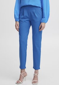 Pantalons bleus en tissu doux, avec un design ajusté comportant des plis devant, une coupe fuselée et une longueur cheville, associés à des talons blancs à lanières.