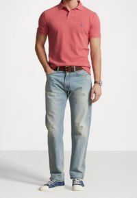 Rode polo shirt met een klein blauw logo, lichtblauwe versleten jeans, bruine riem en blauwe sneakers met witte zolen. Over het algemeen een casual outfit.