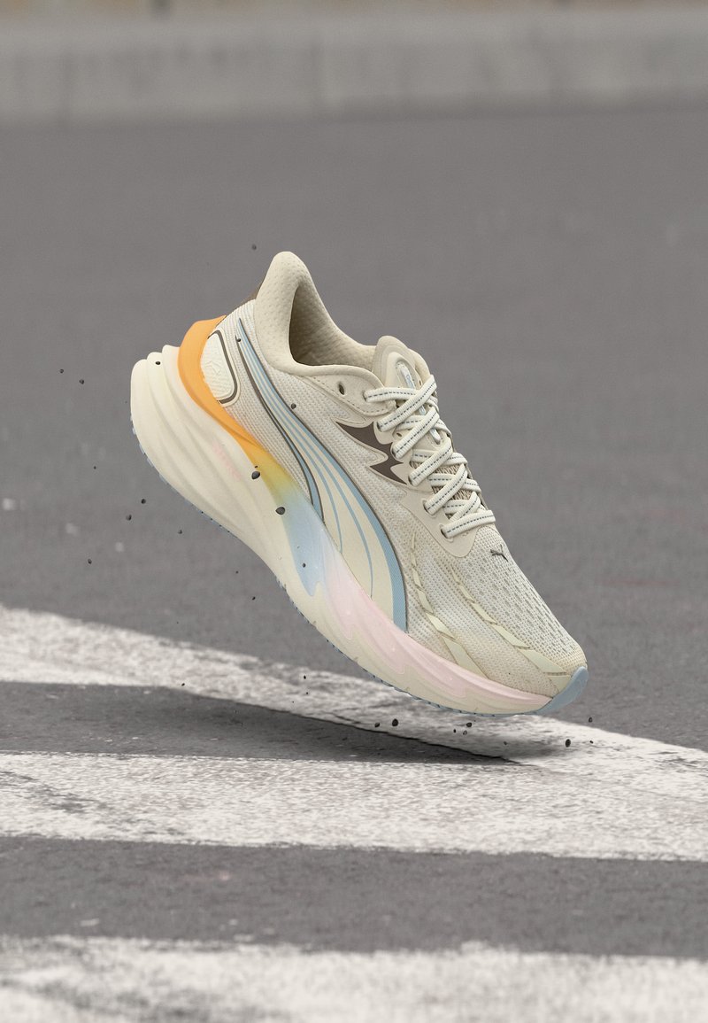 Ανοιχτό μπεζ αθλητικό παπούτσι με παστέλ μπλε και πορτοκαλί λεπτομέρειες, αιωρούμενο πάνω από έναν ασφαλτοστρωμένο δρόμο με λευκές βαμμένες ρίγες και μικρά απορρίμματα.