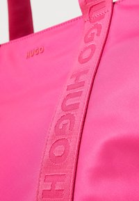 Rozā auduma iepirkumu soma ar gludu tekstūru un uzspiestu "HUGO" logo uz siksnas. Ietver platu siksnu ērtai nēsāšanai.