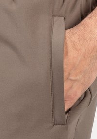 Braunes Stofftaschendetail mit sichtbaren Nähten. Glatte Textur, taillierte Passform und ein teilweise in die Tasche gesteckter handfarbiger Hand.