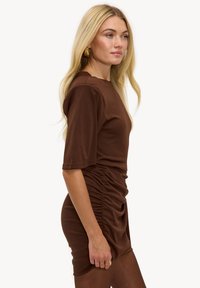 Bruine bodyconjurk gemaakt van zachte stof; heeft korte mouwen, een gedetailleerde geplooide zijkant en een gladde textuur.