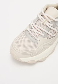 Beige sportcipő, hálós felsőrésszel és szintetikus betétekkel. Jellemzői a textúrált felület, kerek orr és vastag gumitalp. A fűzők azonos színűek.