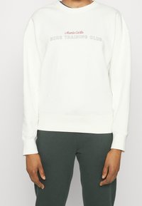 Persona che indossa un felpa bianca con "Monte Carlo Borg Training Club" sul petto e pantaloni verde scuro, in piedi contro uno sfondo neutro.