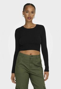 Fekete bordás, hosszú ujjú crop top, olívazöld cargo nadrággal kombinálva, amely zsebekkel és gombos záródással rendelkezik.