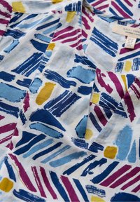 Tessuto in cotone biologico caratterizzato da un vivace pattern astratto con disegni a pennellate in blu, viola, giallo e bianco. Dettaglio a V visibile.