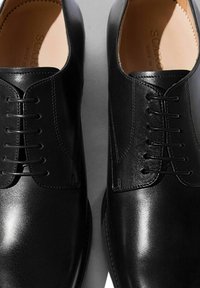 Scarpe eleganti in pelle nera con texture liscia, caratterizzate da una punta appuntita, un design con lacci a sette occhielli e dettagli di cucitura sottili.