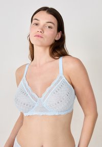 Bralette en dentelle bleu clair avec des motifs circulaires complexes, des bords en festons et des bretelles réglables. Texture douce et transparente avec une forme triangulaire.