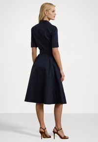 Navy blue dress with short sleeves, button-down collar, and cinched waist. Flared skirt with pleats, made of smooth fabric. Brown heeled sandals.
